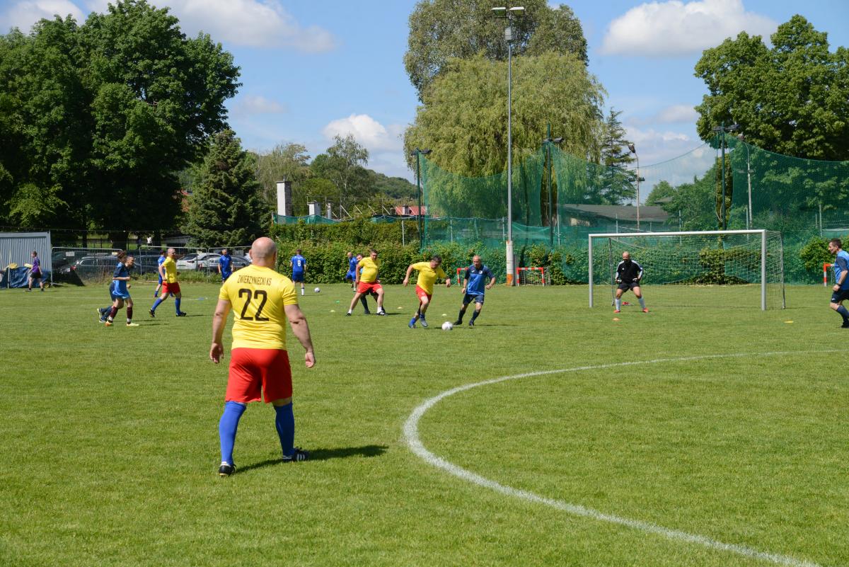 Zdjęcie nr: 10-DSC_3244 przedstawia odbywający się mecz piłkarski drużyn ubranych w żółtoczerwone stroje oraz w stroje granatowe. W bramce stoi bramkarz ubrany w czarny strój. Dwóch mężczyzn kopie piłkę.