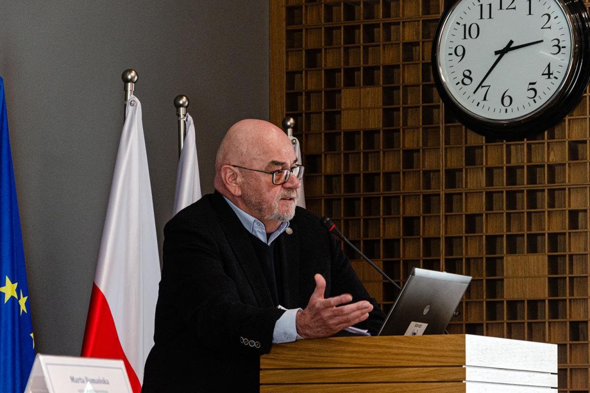 Przedstawia mężczyznę prof. dr. hab. Jerzego Zajadło. Mężczyzna w okularach, ubrany w czarną marynarkę i błękitną koszulę stoi za mównicą i przemawia. Za nim widać flagę państwową oraz dwa białe wierzchołki innych flag. Mężczyzna jest ujęty z profilu nad nim, na drewnianej ścianie wisi duży, biały, okrągły zegar. 