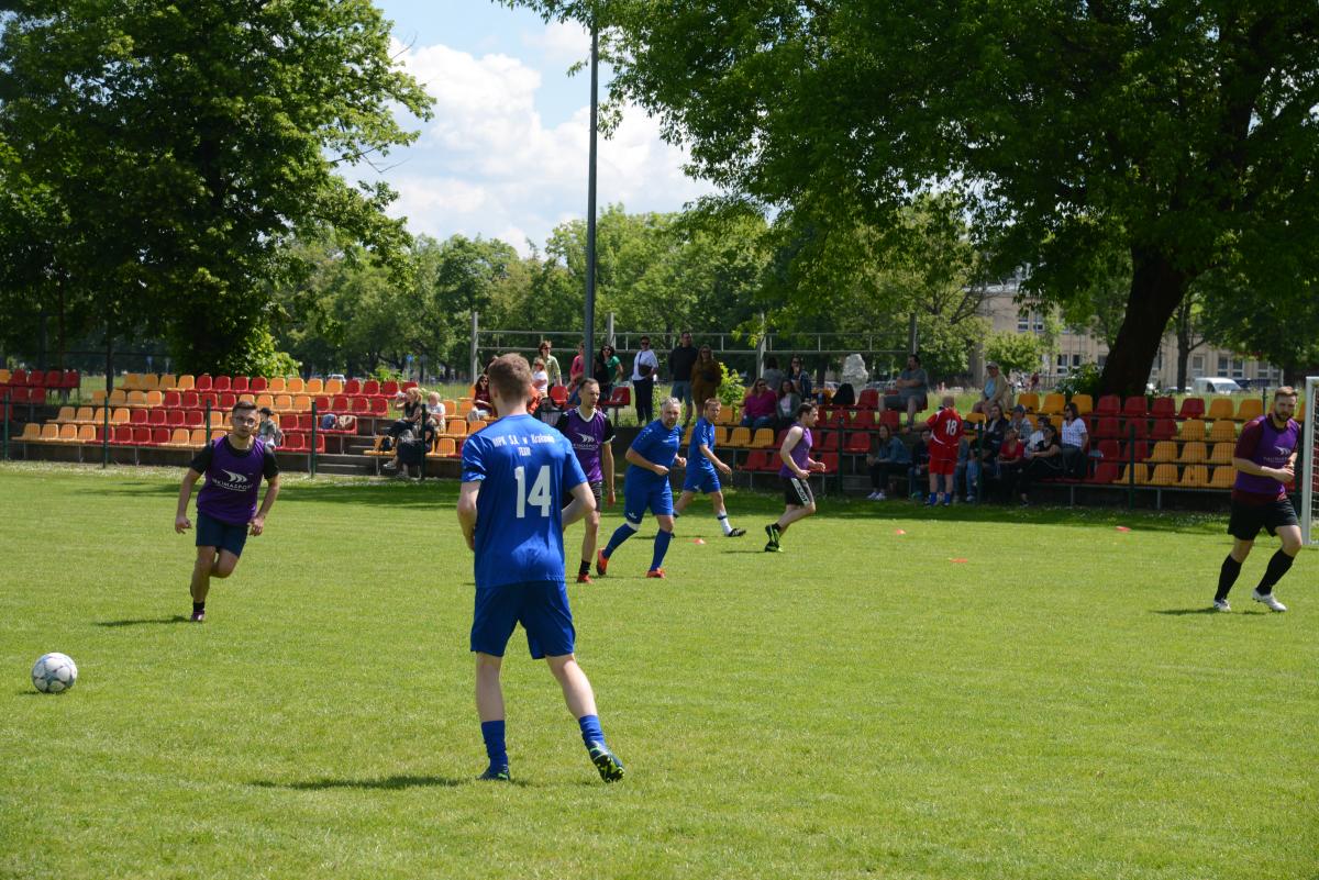 Zdjęcie nr: 17-DSC_3277 przedstawia odbywający się na murawie mecz piłkarski drużyn ubranych w niebieskie  stroje oraz w stroje fioletowoczarne. Zawodnicy biegają po boisku. W tle trybuny z publicznością – kibicami.