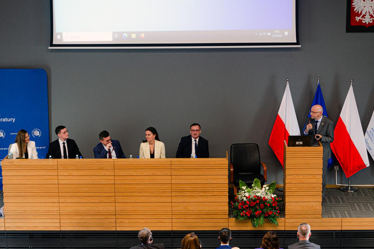 Zdjęcie z panelu dyskusyjnego, przedstawia od lewej strony siedzących za katedrą kobietę w okularach i białym żakiecie, aplikantkę aplikacji prokuratorskiej, Panią Magdalenę Szczurek, za którą znajduje się fragment niebieskiej ścianki reklamowej Krajowej Szkoły Sądownictwa i Prokuratury. Obok niej znajduje się mężczyzna w okularach, czarnej marynarce i krawacie oraz białej koszuli, aplikant aplikacji sędziowskiej, Pan Konstanty Kuryłowicz, następnie pochylony mężczyzna w okularach, granatowej marynarce, białej koszuli i bordowym krawacie, Pan prof. Uniwersytetu Śląskiego dr hab. Krystian Markiewicz – Prezes Stowarzyszenia Sędziów Polskich Iustitia. Następnie widać kobietę w kremowym żakiecie i czarnej bluzce, Panią adwokat Paulinę Kierzkowską-Knapik oraz mężczyznę w okularach, czarnej marynarce i krawacie oraz białej koszuli, Pana Prokuratora Regionalnego w Krakowie Mariusza Krasonia. Nad katedrą widać fragment ekranu do rzutnika, a poniżej biało-czerwoną wiązankę kwiatów, za którą stoi krzesło. Za mównicą stoi mężczyzna w okularach i szarym garniturze, Dyrektor Krajowej Szkoły Sądownictwa i Prokuratury, prof. dr hab. Piotr Girdwoyń, który trzyma mikrofon zamontowany w mównicy. Przed nim stoi laptop. Za nim znajdują się flagi, kolejno od lewej: państwowa, unijna, państwowa. Widoczny jest fragment flagi KSSiP.  Flagi umieszczone są na stojakach flagowych. Nad nimi, na szarej ścianie zawieszone jest godło państwowe. Poniżej podwyższenia, na którym znajduje się katedra i mównica widocznych jest pięć osób z publiczności odwróconych tyłem. Zdjęcie wykonano na wprost. 