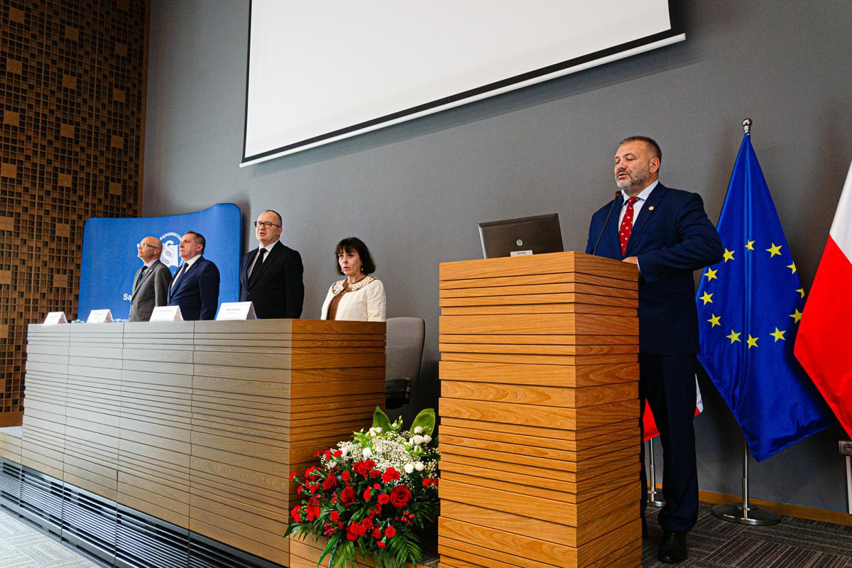 Przedstawia pięć osób – od lewej - Dyrektora Krajowej Szkoły Sądownictwa i Prokuratury, prof. dr. hab. Piotra Girdwoynia, w okularach i ubranego w szary garnitur; zastępcę Prokuratora Krajowego, pana Marka Jamrogowicza, ubranego w granatowy garnitur; Ministra Sprawiedliwości, Pana Adama Bodnara, w okularach i ubranego w czarny garnitur; sędzię Sądu Najwyższego, Panią Martę Romańską, ubraną w kremowy żakiet i brązową bluzkę. Stoją oni za katedrą. Osoby widać tylko do pasa. Obok katedry za mównicą stoi Dyrektor ds. Organizacyjnych, Sędzia Waldemar Żurek. Zdjęcie przedstawia chwilę  z odśpiewania hymnu państwowego. Pomiędzy katedrą, a mównicą znajduje się na podłodze biało-czerwona wiązanka kwiatów. Za Dyrektorem Żurkiem znajdują się trzy flagi – dwie polskie, a pomiędzy nimi flaga Unii Europejskiej umieszczone na stojakach flagowych. Nad osobami  znajdującymi się na zdjęciu widać fragment ekranu do rzutnika,  a za dyrektorem Girdwoyniem i panem Jamrogowiczem znajduje się ścianka reklamowa w kolorze niebieskiemu z logo Krajowej Szkoły Sądownictwa i Prokuratury w białym kolorze.  