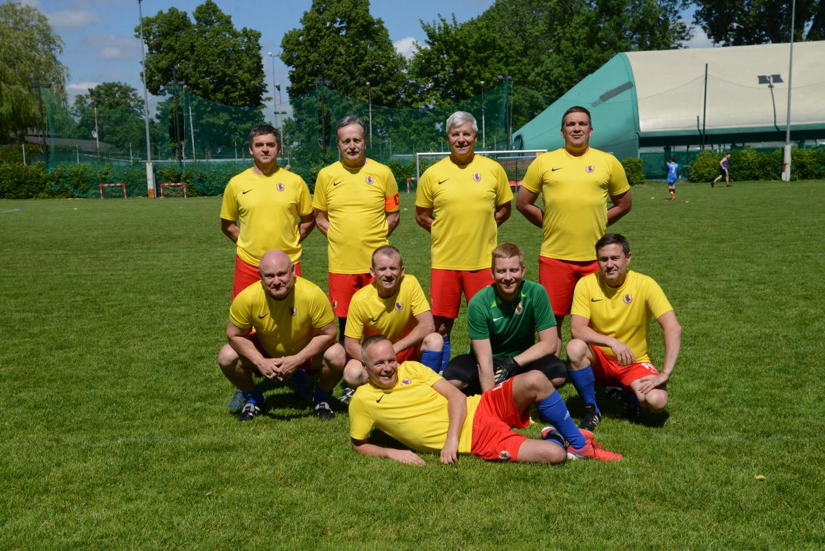 Zdjęcie nr: 5-drużyna przedstawia skład męskiej drużyny piłkarskiej, składającej się z dziewięciu zawodników, na murawie piłkarskiej, ośmiu  z nich ubranych jest w żółtoczerwone stroje a jeden w strój bramkarski koloru zielonego. Czterech mężczyzn stoi, pozostali spoczywają na trawie (czterech kuca, jeden leży).