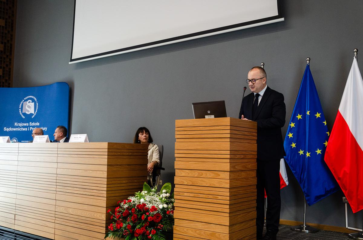 Przedstawia mężczyznę w okularach, czarnym garniturze i krawacie, a także białej koszuli – Ministerstwa Sprawiedliwości, Adama Bodnara. Mężczyzna znajduje się za drewnianą mównicą w naturalnym kolorze jasnego drewna. Przed nim na mównicy stoi laptop, za nim znajdują się dwie flagi narodowe, a pomiędzy nimi flaga Unii Europejskiej, umieszczone na stojakach flagowych. Flagi znajdują się na krawędzi zdjęcia, z drugiej strony stoi ścianka w kolorze niebieskim z białym logo Krajowej Szkoły Sądownictwa i Prokuratury. Nad mężczyzną widać fragment ekranu do rzutnika. Zdjęcie wykonane jest na tle szarej ściany. Obok mównicy widać katedrę, przy której siedzą Sędzia Sądu Najwyższego Marta Romańska, zastępca Prokuratora Krajowego Marek Jamrogowicz oraz Dyrektor Krajowej Szkoły Sądownictwa i Prokuratury Piotr Girdwoyń. Pomiędzy katedrą, a mównicą  znajduje się wiązanka z biało-czerwonych kwiatów. 