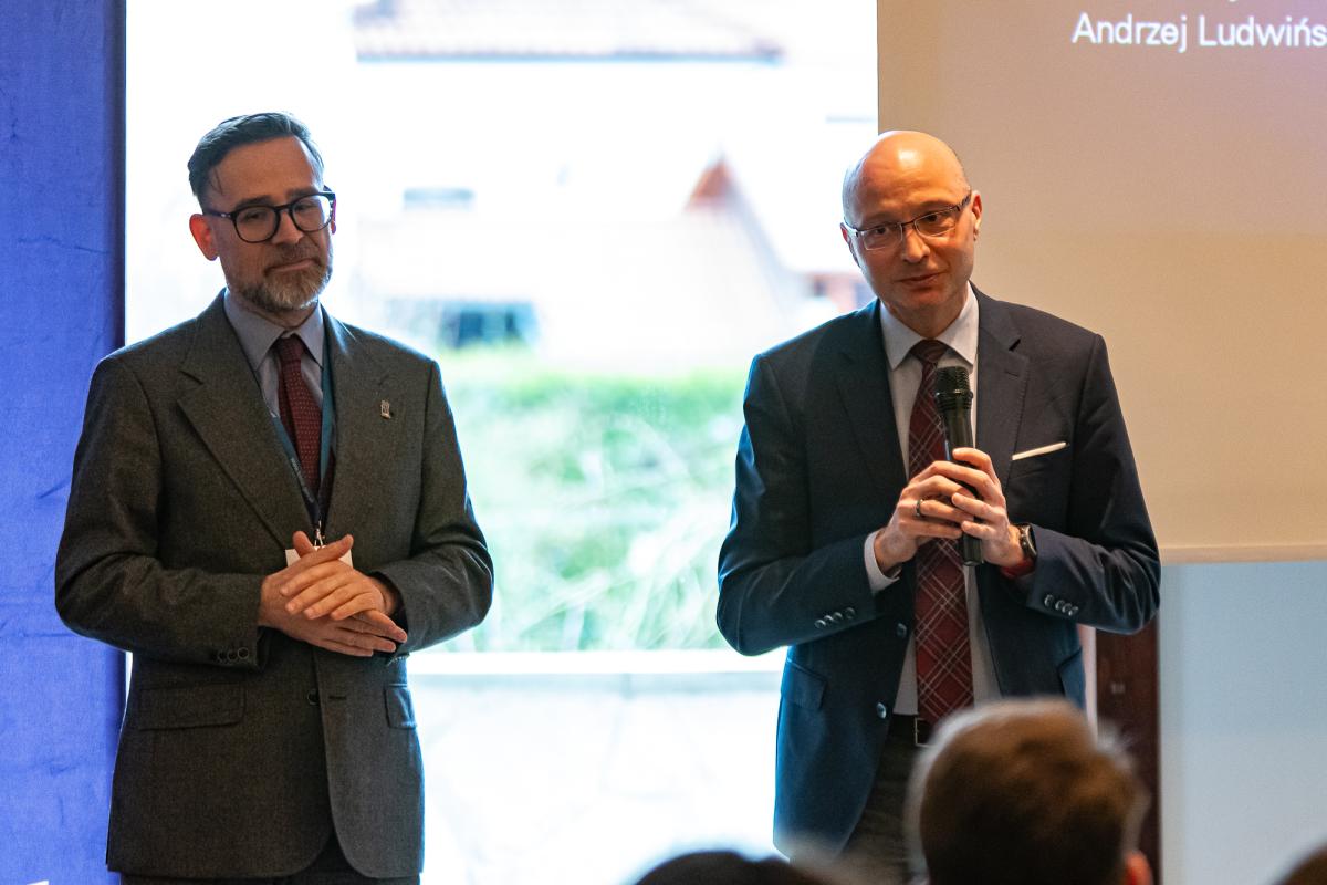  dwóch mężczyzn stojących przez publicznością – uczestnikami konferencji, jeden z mężczyzn trzyma mikrofon w ręce i przemawia do zgormadzonych ludzi