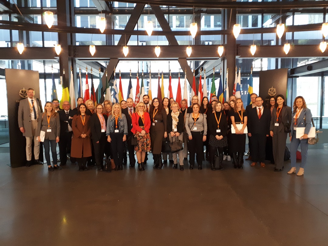 Uczestnicy wydarzenia pozują do grupowego zdjęcia stoją w budynku Trybunału Sprawiedliwości Unii Europejskiej w tle flagi narodowe krajów UE