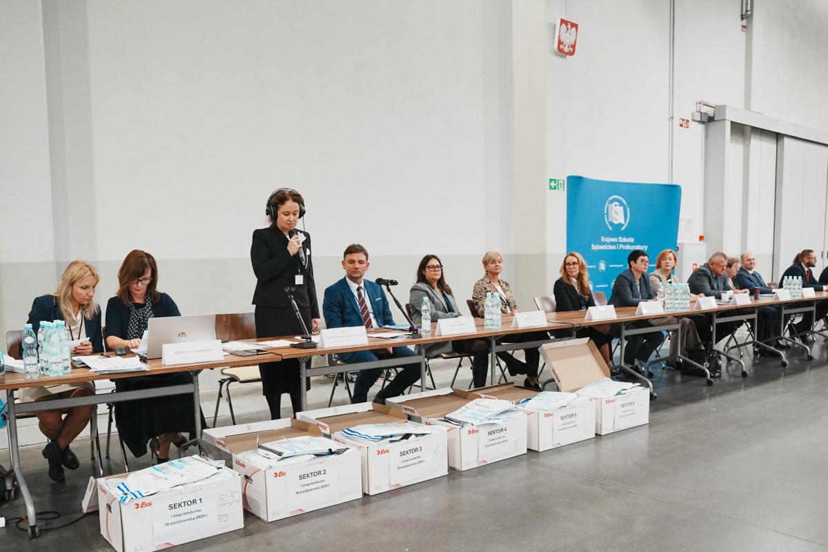 kobieta z mikrofonem w ręce stoi za stołem i przemawia do uczestników konkursu, obok siedzi osiem kobiet i pięciu mężczyzn za stołem. Przed sobą mają na stole ustawione tabliczki z imieniem i nazwiskiem. Za ich plecami ustawiony jest baner w kolorze biało-niebieskim z napisem Krajowa Szkoła Sądownictwa i Prokuratury. Przed stołem leży sześć pudeł z zapakowanymi w koperty bezpieczne materiałami na konkurs.